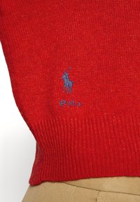 Punainen neulottu villapaita, jonka helmassa on sininen brodeerattu Polo-logo hevosesta ja ratsastajasta, yhdistettynä beigeihin housuihin.