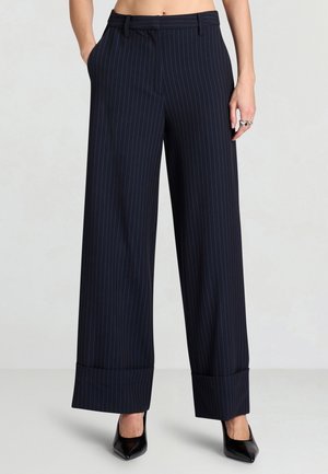 Femme portant un pantalon large à rayures bleu marine avec revers et des talons pointus noirs, les mains dans les poches.