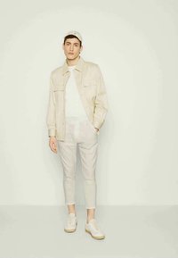 Chaqueta de camisa beige clara con dos bolsillos frontales, combinada con camiseta blanca y pantalones cortos beige claros. Zapatillas blancas con detalles beige.