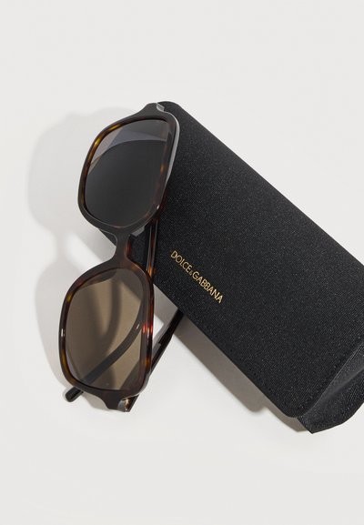 Γυαλιά ηλίου με σκελετό σε σχέδιο χελώνας και σκουρόχρωμους φακούς, τοποθετημένα πάνω σε μαύρη υφασμάτινη θήκη Dolce & Gabbana σε ανοιχτό φόντο.