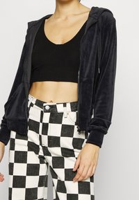 Femme portant un haut court noir, une veste à zip noire et un pantalon à carreaux noir et blanc, les mains détendues sur les côtés.