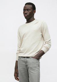 Άνδρας που φοράει μπεζ μακρυμάνικη μπλούζα και ανοιχτό γκρι τζιν, στέκεται με το ένα χέρι στην τσέπη του μπροστά από λευκό φόντο.