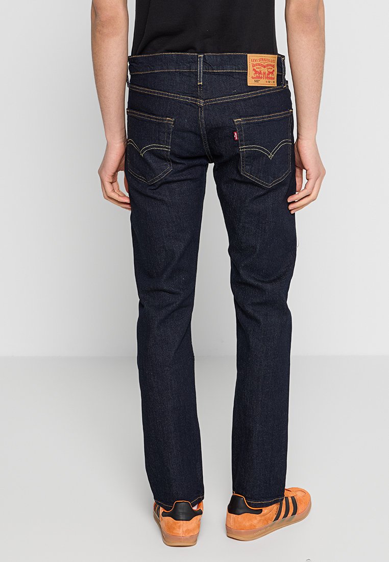 Jean en denim bleu foncé avec des coutures jaunes, coupe droite, poches arrière visibles et étiquette en cuir à la taille. Porté avec des baskets oranges.