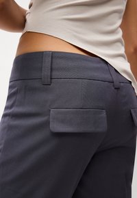 Pantaloni grigi scuri con una finitura testurizzata, dotati di due tasche quadrate posteriori e di un girovita piatto, parzialmente visibili sotto una maglietta di colore chiaro.