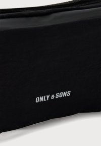 Черна ткан с бял надпис "ONLY & SONS" отпечатан близо до долния ръб, показващ част от облекло или аксесоар.