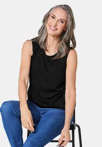 Top nero senza maniche con scollatura rotonda, tessuto liscio e vestibilità comoda, abbinato a jeans blu, seduto su una sedia nera.