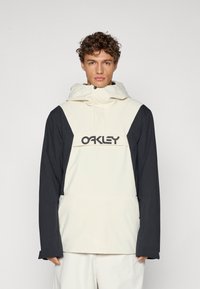 Cremefarvet og sort hættetrøje med et logo på forsiden, fremstillet af let materiale, løs pasform og lange ærmer med justerbare manchetter.