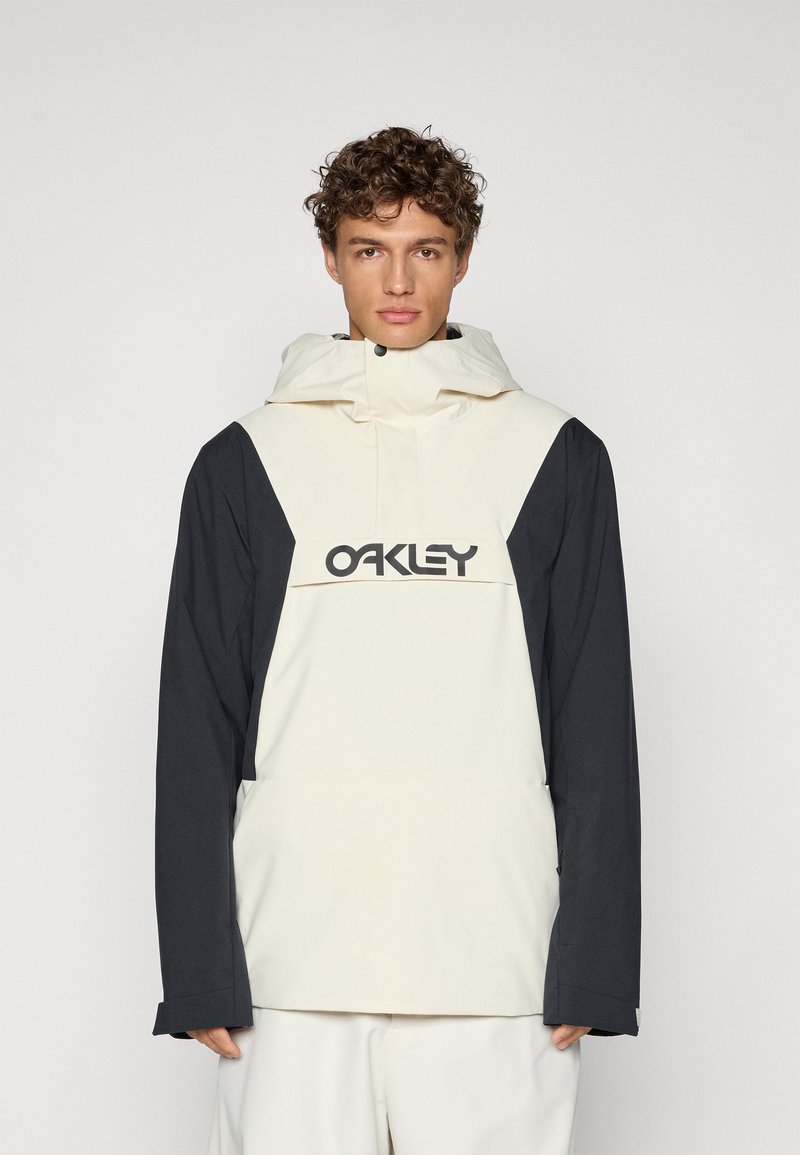 Veste à capuche crème et noire avec un logo à l'avant, fabriquée dans un matériau léger, à coupe ample et avec des manches longues à poignets ajustables.