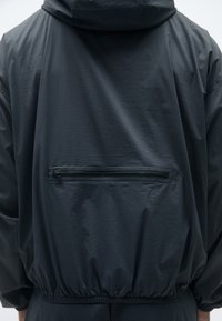 Veste légère gris foncé avec capuche, poignets élastiques et une poche zippée distincte dans le dos. Texture lisse avec un léger éclat.