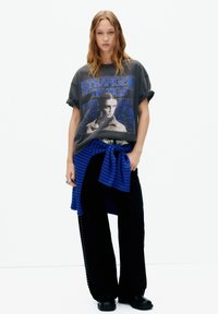 Tricou gri grafic cu imprimeu albastru "Stranger Things", asortat cu pantaloni negri cu croială largă și un pulover albastru în dungi legat în talie. Pantofi negri.