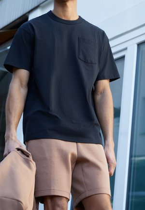 Homme portant une chemise noire à manches courtes et un short beige, tenant un sac beige, debout dehors près de fenêtres en verre.