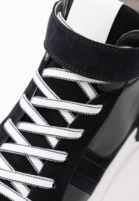 Korkeajalkaiset lenkkarit tummansinisen mokkanahan ja tumman nahan yhdistelmässä, joissa on valkoiset nauhat ja ompeleet sekä pehmustettu nilkkaremmi tuen tarjoamiseksi.