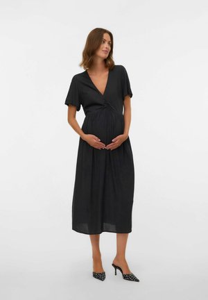 Femme enceinte en robe midi noire à manches courtes, tenant son ventre, portant des talons pointus noirs à pois blancs.