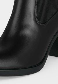 Bottine noire en cuir avec un bout arrondi, un panneau latéral élastique et un talon bloc sur fond blanc.
