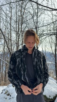 Sort og grå fleecejakke med lynlås og et tekstureret, abstrakt mønster. Har en høj krave og elastiske manchetter, båret over en mørk skjorte.