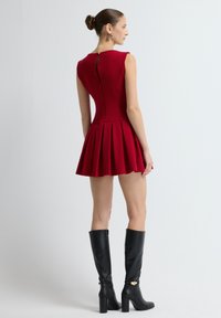 Mujer con el cabello recogido en un moño, vistiendo un vestido mini sin mangas de pliegues rojos y unas botas negras de tacón alto hasta la rodilla, de pie frente a un fondo blanco liso.