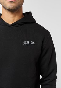 Muž v černé mikině s kapucí s bílým vyšitým textem „CLSR LDN Design Atelier“ na levé straně hrudi, neutrální šedé pozadí.