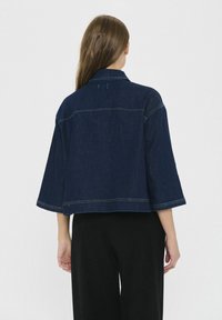 Ženska oblečena v temno modro denim jakno s širokimi rokavi, gledano s hrbta, v kombinaciji s črnimi hlačami.