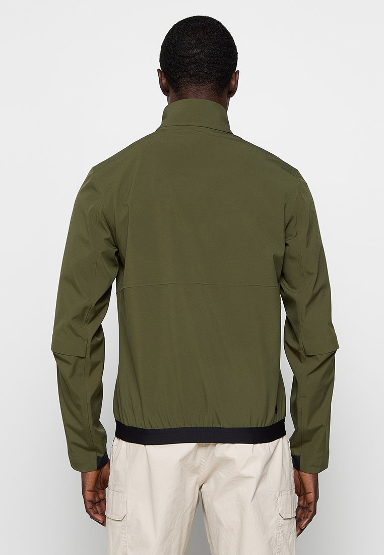 Giacca impermeabile verde oliva con colletto alto, polsini elasticizzati e orlo nero. Presenta un tessuto liscio e leggero e un design minimalista.