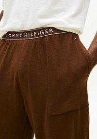 Primo piano di pantaloni marroni Tommy Hilfiger con vita elastica e scritte bianche, indossati con una camicia bianca, mano in tasca.