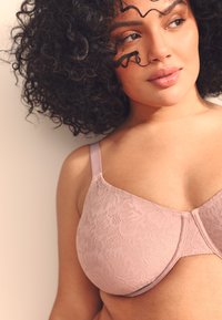 Un soutien-gorge en dentelle rose clair avec un motif floral, des bretelles réglables et une texture lisse. Les bonnets ont une forme arrondie avec des coutures discrètes.