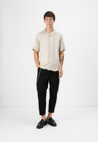 Short-sleeve beige shirt with a casual collar, black tapered pants, and black loafers. Features a chain accent on the pants.