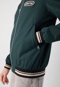 Personne portant une veste bomber vert foncé avec des poignets et une ceinture rayés, main dans la poche latérale zippée.