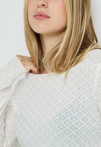 Top bianco a maglia testurizzata con motivo geometrico, vestibilità ampia e maniche larghe. Il tessuto sembra leggero e leggermente trasparente.
