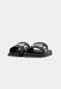 Sorte slidesandaler med brede stropper, der har "HUGO" i lyseblå bogstaver, lavet af gummi, med tekstureret sål og kontrasterende tekstureret bund.