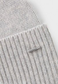 Světle šedá pletená čepice s ribovaným vzorem a stříbrnou kovovou logem. Měkká textura, zaoblený tvar a těsné padnutí.