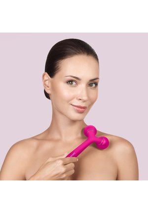 Femme à la peau nette utilise un masseur double tête rose vif sur son épaule sur un fond violet clair uni.