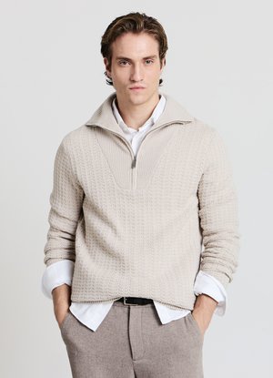 Cremfarbener Strickpullover mit hohem Kragen, ausgestattet mit einem Viertel-Reißverschluss und strukturiertem Muster. Getragen über einem weißen Hemd und hellen Hosen.