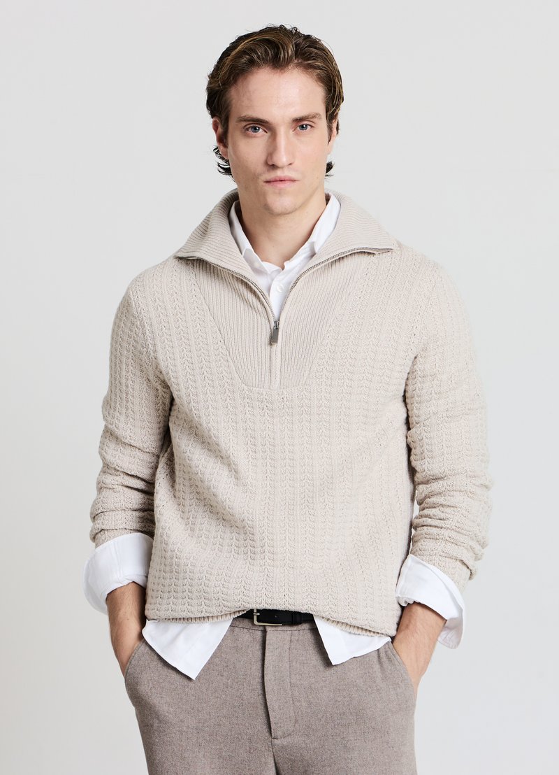Pull en maille crème avec un col montant, présentant une fermeture éclair quart de zip à l'avant et un motif texturé. Porté par-dessus une chemise blanche et un pantalon clair.