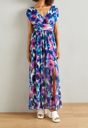 Vestido largo con escote en V, mangas de capa, estampado floral vibrante en azul, rosa y verde, cintura entallada y abertura lateral. Zapatos plataforma blancos.