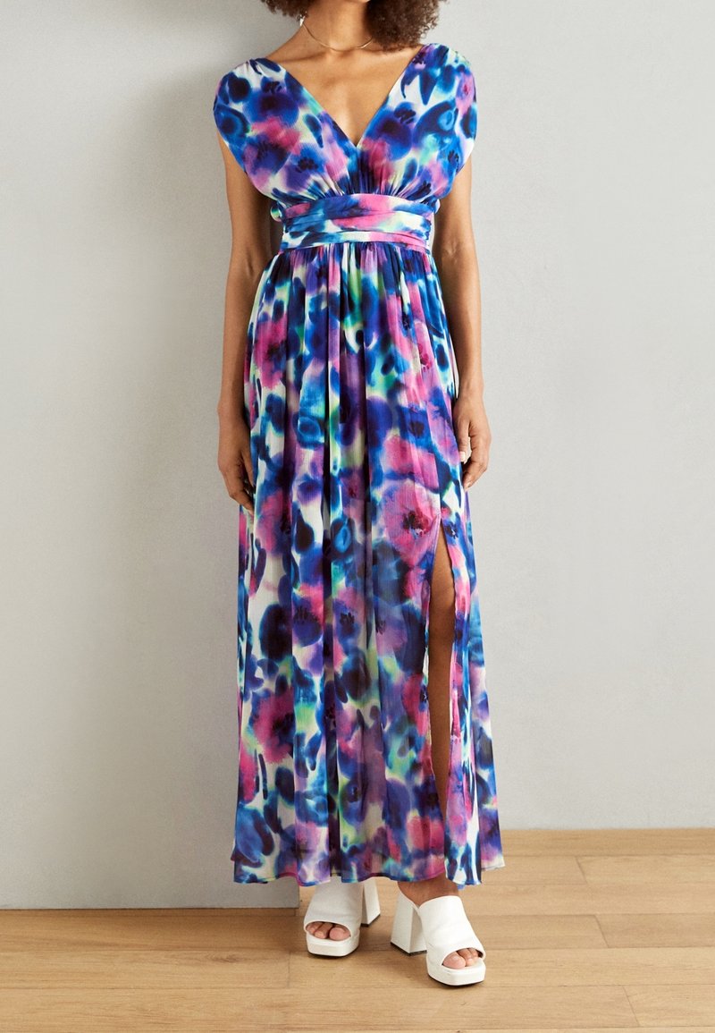 Robe longue avec décolleté en V, manches courtes, motif floral vibrant bleu, rose et vert, taille cintrée et fente latérale. Chaussures compensées blanches.