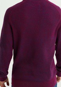 Suéter de punto con cuello redondo en púrpura oscuro, con textura de canalé, que presenta mangas largas y un ajuste relajado. Sin patrones ni acentos visibles.