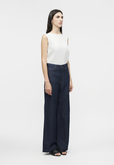 Γυναίκα που στέκεται, φοράει αμάνικη λευκή μπλούζα, ψηλόμεσα φαρδιά σκούρα μπλε τζιν και μαύρα σανδάλια με ανοιχτά δάχτυλα, σε απλό φόντο.