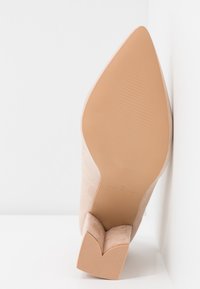 Chaussure beige en suède à bout pointu avec un talon bloc de forme unique, semelle tournée vers l'avant sur fond blanc.