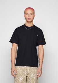 T-shirt nera in cotone a maniche corte con taschino sul petto, caratterizzata da un piccolo logo sulla tasca. Indossata con pantaloni corti beige a fantasia.