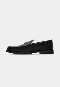 Mocassin en cuir noir avec détail boucle dorée, bout arrondi, dessus cousu et semelle à crampons sur fond blanc.