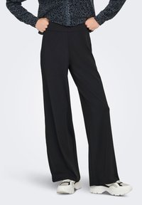 Άτομο με μαύρο παντελόνι φαρδύ στο πόδι, λευκά και μαύρα αθλητικά παπούτσια, και σκούρο ζακέτα με φερμουάρ και σχέδιο, με τα χέρια στους γοφούς.