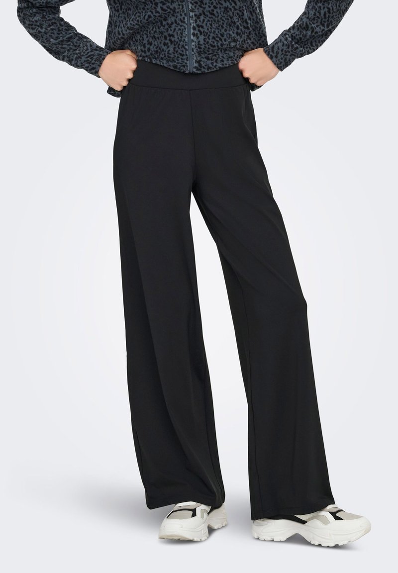 Άτομο με μαύρο παντελόνι φαρδύ στο πόδι, λευκά και μαύρα αθλητικά παπούτσια, και σκούρο ζακέτα με φερμουάρ και σχέδιο, με τα χέρια στους γοφούς.