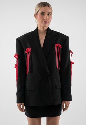 Kobieta o blond włosach nosi oversize czarną marynarkę ozdobioną wieloma czerwonymi kokardami na rękawach i z przodu, zestawioną z czarną spódnicą.