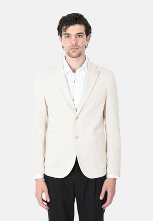 Uomo che indossa un blazer beige chiaro sopra una camicia bianca e pantaloni neri, in piedi contro uno sfondo bianco uniforme.