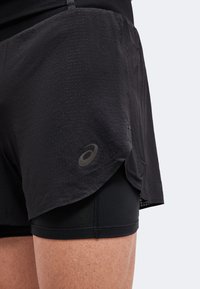 Gros plan d'une personne portant un short de course noir superposé avec un logo ASICS sur la couche extérieure, montrant la partie supérieure de la cuisse et un tissu texturé.
