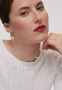 Srebrny naszyjnik z małymi koralikami, z wyraźnym detalem w postaci perły. Modelka nosi prążkowany biały sweter i wiszące kolczyki z perłami.