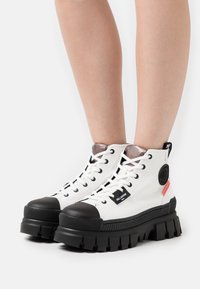 Botas de tobillo de lona blancas con punteras de goma negra y suelas gruesas negras. Presentan un diseño de cordones y acentos de marca en el lateral.