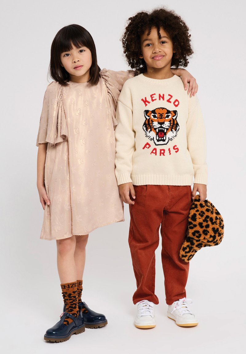 Robe couleur perle avec un tissu texturé et des manches bouffantes ; pull crème avec un graphique de tigre ; pantalon rouille ; chaussures navy ; chapeau et chaussettes à pois.