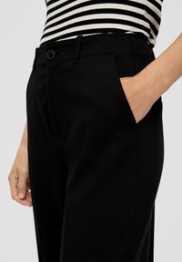 Schwarze, maßgeschneiderte Hose im Slim Fit, mit seitlichen Taschen, glatter Textur und flachem Frontdesign. Getragen mit einem gestreiften Oberteil.