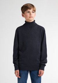 Dunkelblaues Rollkragenpullover aus Strickstoff mit langen Ärmeln und gerippten Bündchen. Kombiniert mit hellblauen Jeans. Glatte Textur.